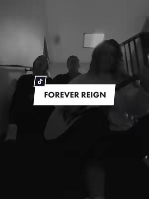 A black and white photo depicts a group of three individuals, the focus is on the middle individual, whose face is not fully visible. They are standing on a staircase or a similar structure. The top of the framing has a white rectangle overlay that contains the text: “FOREVER REIGN”. The TikTok logo can be seen on the top left corner.