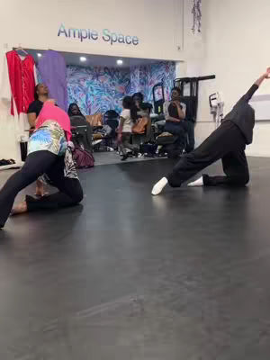 Two people kneeling, both with one leg in front and another in a kneeling position, posing in a dance studio with other people in the background. The person in the foreground is wearing a black top with black pants and sock like shoes, posing to the right and left, the person in the left foreground is wearing black pants with white socks.