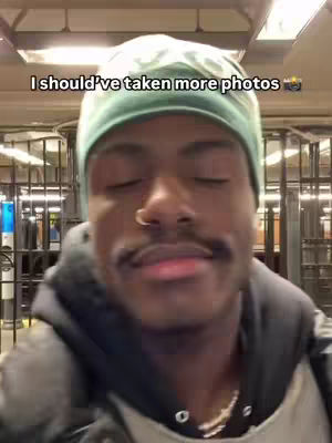 A close-up shot of a young Black man with a green beanie, closed eyes, and a slight smile. He has a mustache and a septum piercing. The background is a blurred urban setting, possibly a subway station or a similar public space. The text overlay reads, 'I should've taken more photos'.