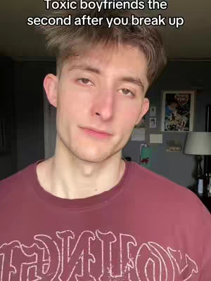 The image shows a close-up of a young man with light skin and short, styled brown hair. He is wearing a maroon t-shirt with a graphic design in white. The background shows part of a room with a dark wall and a few decorations. The man has a subtle expression and a slightly downward gaze.