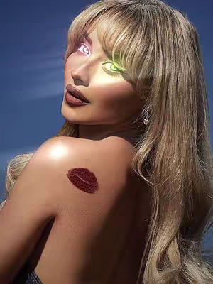 A woman with long blonde hair is captured from the back, looking over her shoulder with a sultry expression. Her eyes are digitally enhanced with glowing white and green effects. She has a dark lip color and a kiss mark on her shoulder. The background is a bright blue sky.