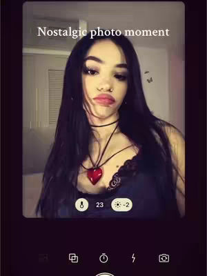 The image shows a young woman posing for a selfie, set against a backdrop that suggests an interior shot. She has long, dark hair and is wearing a black top with a heart-shaped pendant around her neck. Above her, white text reads 'Nostalgic photo moment'. The interface includes camera controls at the bottom, implying the use of a photo-taking app.