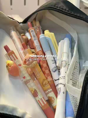 The image shows a close-up view of a pencil case filled with various pens. The pens are of different colors and designs, with some having cartoon characters and decorative patterns. They are packed into a black pencil case, with the zipper partially open. Some pens have accents of light blue and yellow. The background is very blurred, which suggests a shallow depth of field, focusing on the pens while keeping the surrounding area out of focus.