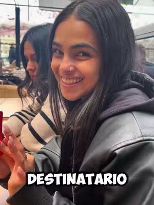 A selfie from the chest up shows two young women. The main subject in the foreground is centered, looking directly at the camera with a cheerful smile, and the woman in the background is smiling and slightly out of focus. The woman in the foreground has long, dark hair and a black leather jacket over a hoodie. The background appears to be a setting indoors. The text overlayed on the bottom reads DESTINATARIO.