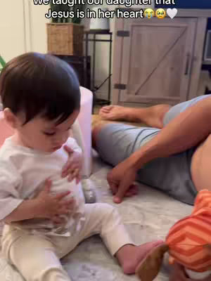 A young child sits on a floor, looking towards their stomach. They are wearing a white shirt and pants. A person's arm and legs can be seen in the background, along with a wooden cabinet, a toy and a woven basket can be seen in the background. Text overlayed states "We taught our daughter that Jesus is in her heart"