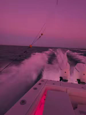 A fisheye view from the deck of a boat during a sunset. The sky is a gradient of pink and purple. The water splashes out from the back of the boat. The boat's structure is white, with the boat's engine clearly visible.