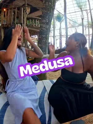 Two women are outside, possibly at a resort. One woman raises her hands toward her face while laughing. She wears a light blue dress. The other woman is to her right and is also laughing. She wears a black top and the word "Medusa" appears overlaid on the image in a rounded purple box.