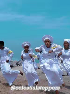 A group of people dressed in white robes and headwear are dancing on a beach. The sky is a clear blue, and the overall lighting is bright. The composition is from a medium angle, capturing the dancers in motion.