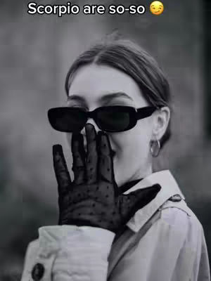 A monochrome image of a woman is shown from the chest up. She wears dark sunglasses and a black, dotted lace glove, her other hand covering her mouth. The background is blurred, emphasizing the subject. The overall composition is a portrait.