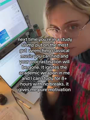 A selfie-style video shows a person with glasses sitting in front of a computer screen and laptop, both displaying content. The person has blonde hair and looks towards the camera. Text overlays appear on the image, describing a study tip with classical music.