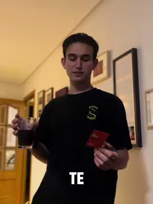A man holding a glass of red liquid and a red card, standing in a hallway with framed pictures on the wall. The text "TE" is written over the image.