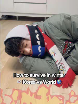 A person is lying on the floor, wrapped in a jacket, hoodie, and scarf, appearing to be sleeping. The scarf is red and black with white text. The floor has a wood-like texture, and there is a cabinet in the background. Text overlayed says, "How to survive in winter Korea vs World".