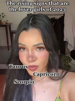 A selfie-style video featuring a young woman with long dark hair. She's wearing makeup, including blush and lip color, and is positioned in front of a neutral-toned background. Overlay text identifies three zodiac signs: Taurus, Capricorn, and Scorpio.