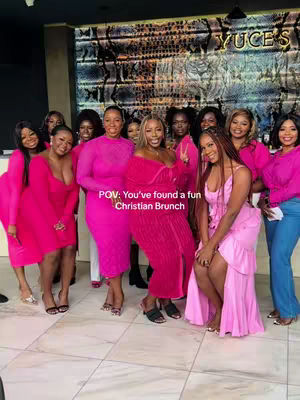 A group of about a dozen women pose for a photo in a polished space. They are all dressed in vibrant pink dresses and tops, some wearing heels, suggesting a social event. The background features a textured wall, a sign with a logo, and a window, with warm lighting enhancing the scene.