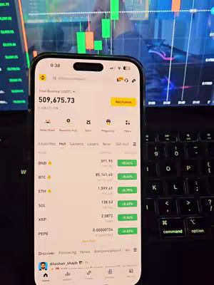 The image showcases a smartphone displaying a trading app interface, held in front of a laptop and a second monitor showing stock charts. The phone's screen prominently features financial data, with a large balance visible at the top. The background includes a laptop keyboard and a blurred-out stock price chart and other details of stock trading, suggesting content focused on trading or financial markets.
