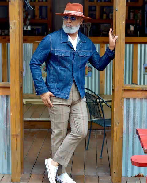 A man in a denim jacket, orange hat, and sunglasses stands in front of a building. He is wearing a white shirt, plaid pants, and white sneakers. The building is made of wood and has a corrugated metal roof. There are plants and other decorations around the entrance.