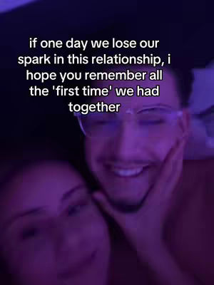 A close-up shot of a couple. The man, wearing glasses, is smiling lovingly at the camera. The woman is partially visible, with her head resting on the man's shoulder. The background is blurred, with a soft violet hue that adds a romantic and intimate feel.