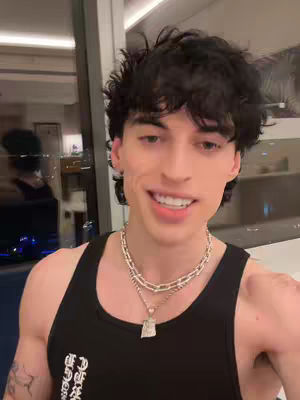 A young man with dark, wavy hair is smiling at the camera. He is wearing a black tank top and two silver chain necklaces. In the background, a reflection of a person can be seen in a glass window, and a bright light.