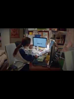 A person with long brown hair sits at a desk with two computer monitors, paperwork, and a cluttered background. They are wearing a blue and white top, facing the monitors as if studying. The lighting is dim, suggesting an indoor setting.