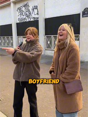 The cover image shows two young women standing outdoors. The woman on the left is holding a microphone and pointing to the right while wearing a brown jacket. The woman on the right is laughing, wearing a long brown coat, and carrying a purse. The background consists of a building facade with graffiti and closed storefront shutters.