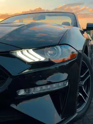 A close-up shot of the front of a black convertible car with its headlights on. The car is positioned on a slight angle, and the background shows a colorful sunset sky.