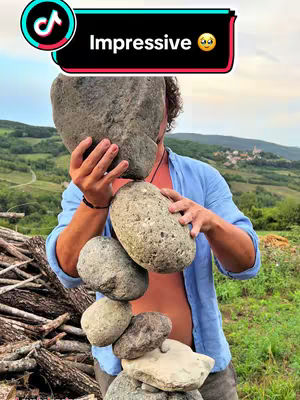 A man balances several stones on top of each other in an outdoor setting. He is wearing a light blue shirt and his face is partially obscured by the stones in front of him. The background features green hills.