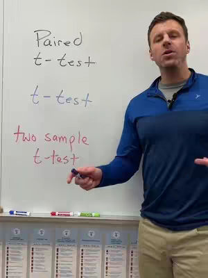 A man stands in front of a whiteboard with statistics concepts written on it. He's wearing a blue and black shirt and tan pants, and is holding a marker.