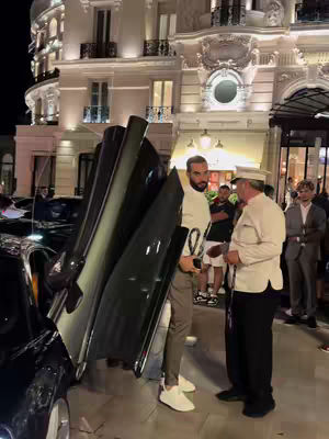 A man exits a black luxury car in front of a grand building at dusk. Other people stand nearby on the sidewalk.