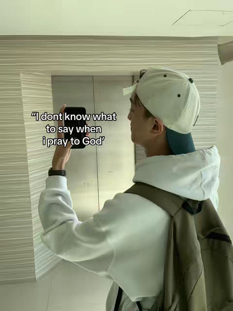 A young man in a white hoodie and a baseball cap is holding a phone to his face, near a paneled wall.
