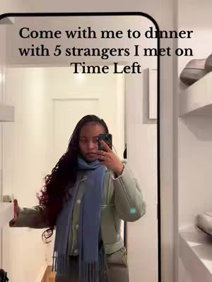 A mirror selfie featuring a woman with long, dark braided hair. She is holding a phone in front of her face, partially obscuring it. She wears a light blue scarf and a green jacket. Text overlayed on top says: "Come with me to dinner with 5 strangers I met on Time Left". The background shows a hallway with a door and white walls.