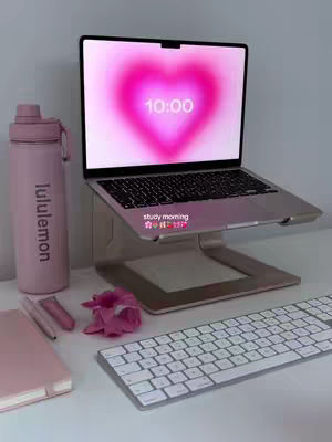 A top-down photo of a workspace with a laptop, water bottle, keyboard, and notebook. The laptop screen displays a heart-shaped gradient at 10:00. The overall aesthetic is pastel pink and white.