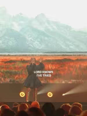 The image shows a live performance with a female singer on stage. The background features a large screen displaying a landscape with mountains and a colorful sky. The singer is wearing a dark dress and is holding a microphone. The lighting is focused on the stage, and there are lights in the foreground.