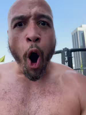 A close-up shot of a bald man with a surprised expression, mouth wide open, with a blurred background featuring a building and a fence.