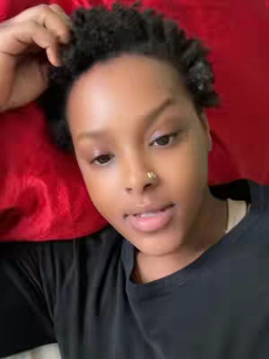 A close-up shot of a person's face lying on a red surface, likely a pillow or bedding. They have brown skin, short, tightly coiled dark hair, and a small nose ring. They are wearing a black top and have a neutral expression, looking toward the camera. The lighting is soft and natural, casting a soft shadow.