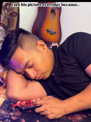 A man is lying on a bed with a black shirt. His head, with short, dark hair, rests on his arm. He is holding a phone. A guitar is in the background.
