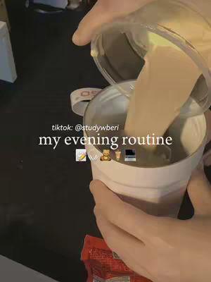 A person is shown pouring a light-colored liquid from a clear container into a white cup. The cup has a label on it, and the person's hands are visible as they perform the action. The background is dark, and there are some other items present.