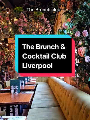 A photograph captures the interior of a stylish restaurant or bar, likely The Brunch & Cocktail Club Liverpool. The space is opulent, featuring plush, mustard-colored seating and tables set for dining. Overhead, vibrant floral arrangements composed of pink flowers and green foliage create a natural, elegant ambiance, with soft lighting illuminating the scene. A large rectangular graphic in the foreground announces "The Brunch & Cocktail Club Liverpool" against a background of teal and black.