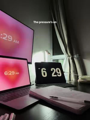 A dimly lit room with a desk is shown, containing a laptop, tablet, and a large monitor, each displaying the time, with a heart graphic on the laptop screen. A notebook and pen are visible. The lighting suggests it is early morning.