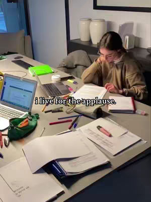 A young woman sits at a desk covered in papers, books, and office supplies, studying in a well-lit room. The image has a warm, inviting tone, with a laptop open in the foreground. A text overlay reads "i live for the applause."