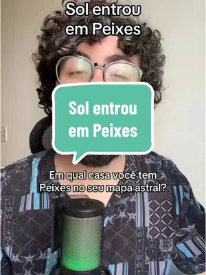 A man with curly dark hair and glasses is centered in the frame, looking towards the camera. The background is a plain, lighter tone. He is wearing an unbuttoned patterned shirt with blue and black colors, and a microphone with a green light is positioned in front of him. Overlaid are speech bubbles with text.