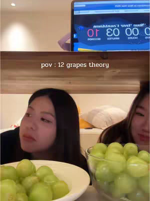 The cover image shows two young women sitting in a room with a wooden wall. They are positioned close to the camera. One woman is looking in the direction of a bowl of grapes with a serious expression. The other woman is looking towards her with a subtle smile. In the background, a laptop displays a timer at the top. The overall composition emphasizes the women and the grapes, suggesting a challenge or game.