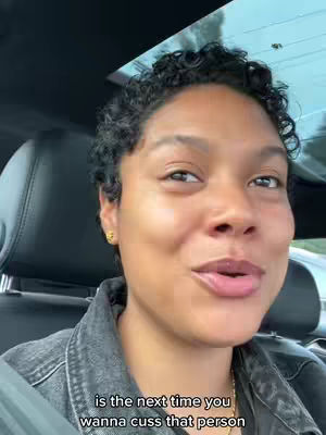 A selfie shot from inside a car. The creator is a woman with short, dark curly hair, wearing a dark denim jacket. She is looking towards the camera and speaking. The background shows the interior of the car, the headrest, and a partially visible window.