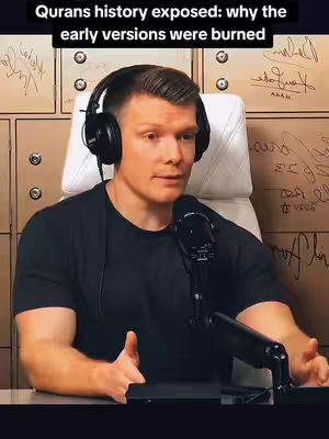 A man with short light brown hair, wearing a black t-shirt and headphones, speaks into a microphone. He is seated in front of a backdrop made of wooden panels with handwriting on them.