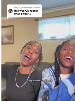The cover image features two young Black women, likely twins, in a casual indoor setting. They are both smiling and have their mouths open as if singing or laughing. The woman on the left is wearing a black top and a silver necklace, while the woman on the right is wearing a blue and white patterned jacket. The background includes glimpses of a cream-colored wall and a window. Text overlay indicates this video is in response to a comment.