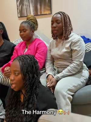 The image shows three Black women seated on a couch, with a partially visible fourth person on the left. The primary focus is on the three women. The woman in front has black braids and is mid-sentence. The woman in the middle is wearing a pink sweater , her eyes closed.. The woman on the right has braided hair and is wearing gray apparel. The background includes a framed picture and a patterned cushion. Overall, the image captures a moment of intimacy and shared experience.