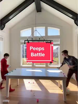 Two young men are in a bright, modern room with a large white table in the center. The text overlay states: Peeps Battle. There is a large window in the background overlooking a suburban landscape, with light-colored walls and a dark ceiling beam. The men appear to be playing a game across the table.
