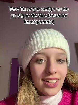 A selfie-style shot of a young woman wearing a white beanie and a pink sweater. The background is slightly blurred, with hints of a purple wall and a decorative object. The woman is smiling, her face filling most of the frame.