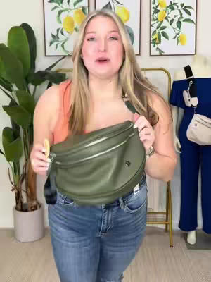 A woman stands in front of a room, holding a green bag, smiling and talking directly at the camera. She is wearing a peach-colored tank top, blue jeans, and has long blonde hair. The background includes a potted plant, two framed pictures with a lemon theme, and a gold clothing rack. A mannequin wearing a blue outfit is positioned on the right.