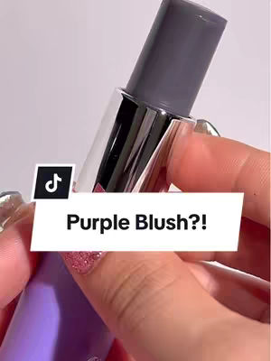 The cover image is a close-up shot focusing on a purple blush stick held in a person's hand, likely a woman. The top of the stick is dark grey, while the rest of the casing is metallic. Her nails are painted with glitter polish. A white, rectangular text box with the words 'Purple Blush?!' in black font is overlaid on the image, with a TikTok logo in the top left corner.