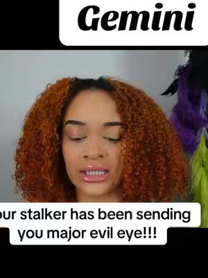 A woman with reddish-brown curly hair and a neutral expression is centered in the frame. The background is a light wall with a purple and green feather boa visible on the right. The top of the image features the word "Gemini". A text overlay at the bottom reads 'Your stalker has been sending you major evil eye!!!'